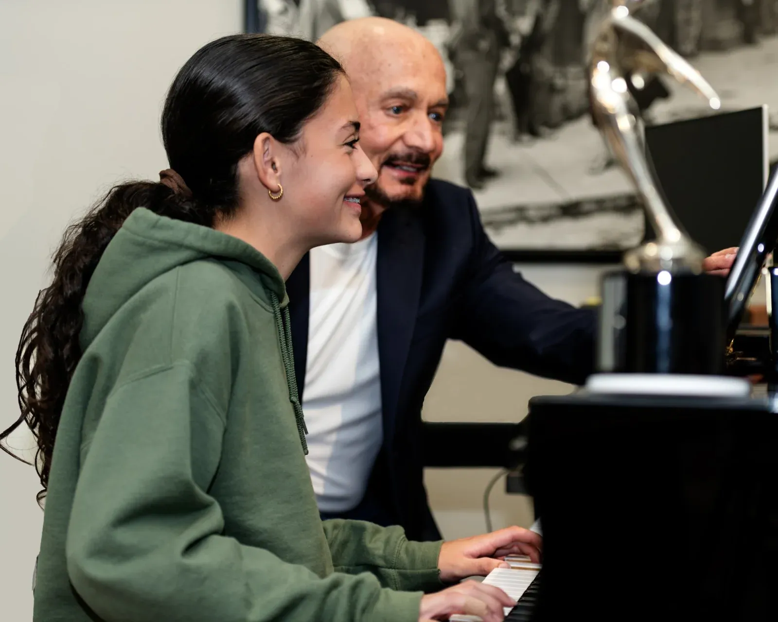 A student smiling as she plays the grand piano, with Thomas leaning in beside her to coach