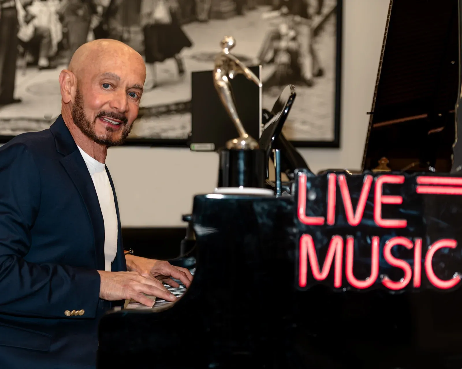 Thomas Calanni smiling at the camera as he plays a black grand piano, a red neon Live Music sign on its lid